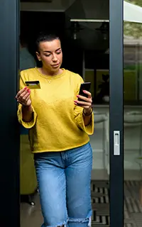 Personne devant une porte vitrée, tenant une carte de crédit et utilisant un téléphone pour un paiement en ligne.