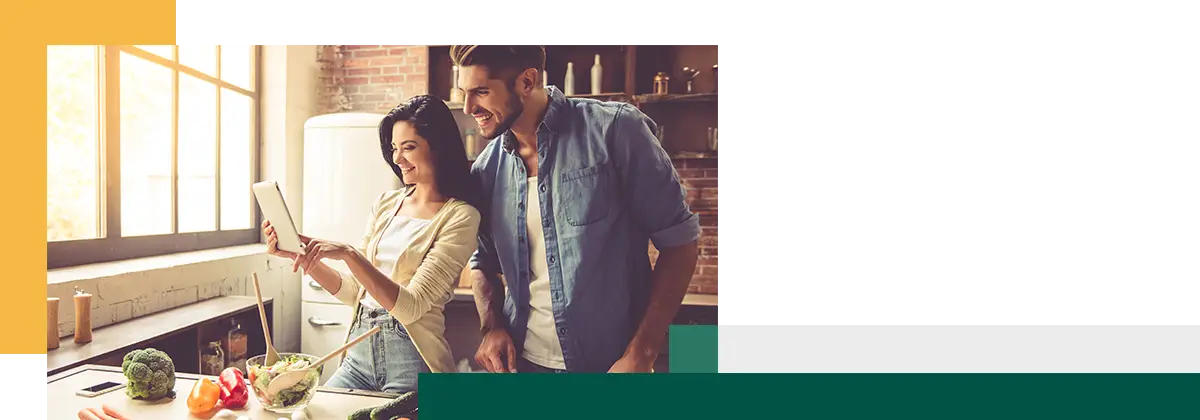 Un jeune couple utilise une tablette numérique et sourit en cuisinant dans leur cuisine.