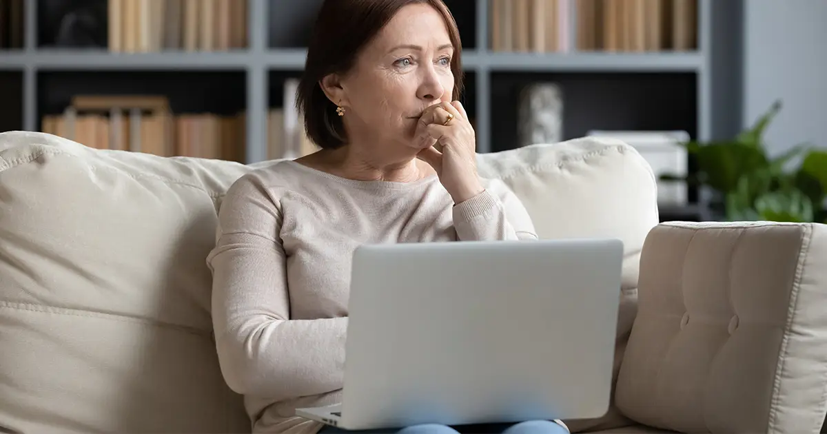 une femme assise sur un canapé dans le salon à l'aide d'un ordinateur portable regarde à distance en pensant ou en réfléchissant