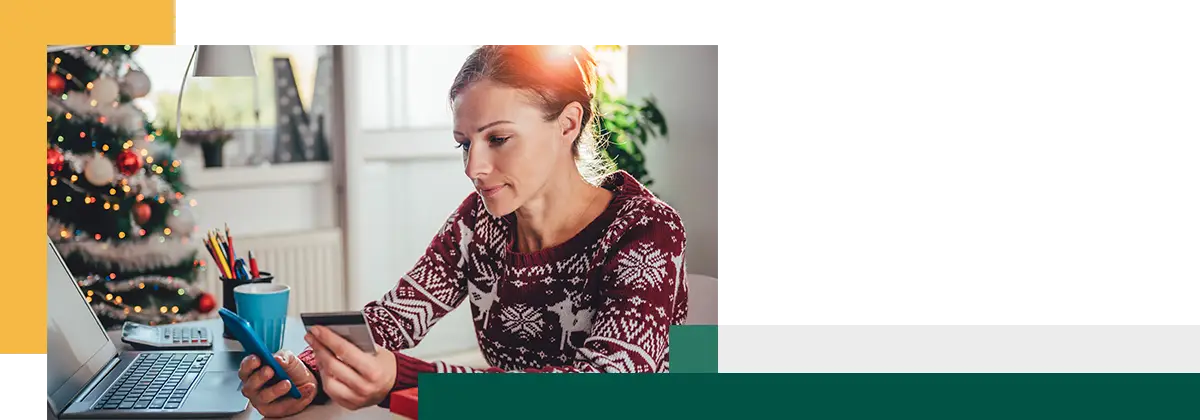 Une femme vêtue d'un pull à motifs est assise à une table, tenant une carte de crédit et une tasse, avec une petite boîte cadeau devant elle.